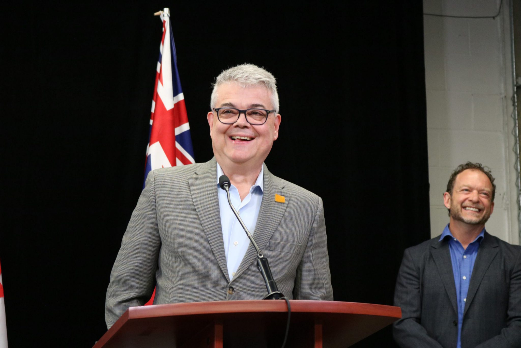 Bill Bradica, Chief Executive Officer, in 2024, smiling while speaking at the podium at a media announcement. Brian Hamilton, 2024 Chair of the Board of Directors, can be seen smiling with Bill in the background.